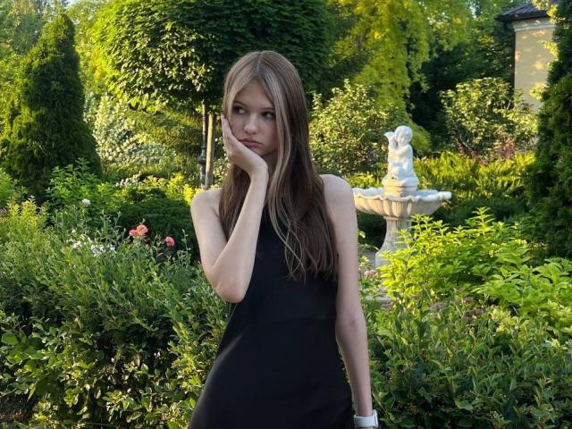SuzieBrion with long brown hair, wearing a black sleeveless dress, touching her cheek with a hand, standing in a lush garden with a white statue in the background.