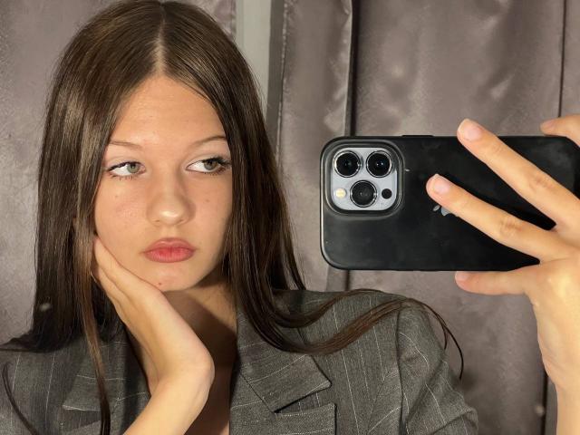 SuzieBrion with long brown hair, pouty pink lips, and a serious expression, holding a black iPhone with three cameras, wearing a gray pinstriped shirt. Her hand's on her cheek.
