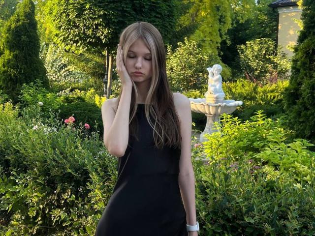 SuzieBrion with long brown hair, wearing a black sleeveless dress, hand on cheek, looking down, green garden with a white fountain in the background, standing with a serious expression.