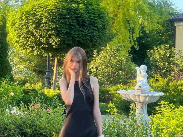 SuzieBrion with long blonde hair, wearing a black sleeveless dress, standing in a garden, hand on cheek, white statue of a cherub on a pedestal to her right.