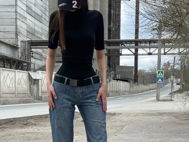 SuzieBrion, wearing a black turtleneck, blue jeans with a black belt, has a slim build, small tits, and long brown hair, standing outdoors by a road.