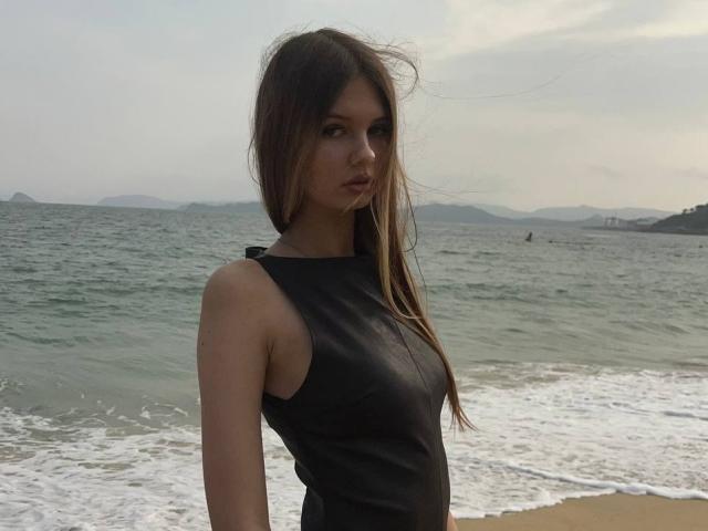 SuzieBrion stands at the beach, wearing a sleeveless black top, with long brown hair and a serious look, near the ocean, showing her tits and ass subtly.