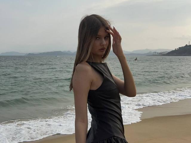 SuzieBrion with long brown hair, wearing a black leather dress, standing on a beach with her hand in her hair, looking at the camera, near ocean waves.
