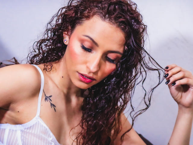AnnaMary, com cabelos castanhos cacheados, blush rosa e delineador preto, vestindo uma blusa branca transparente, mostrando uma pequena tatuagem de uma folha preta no ombro direito, olhando para baixo..