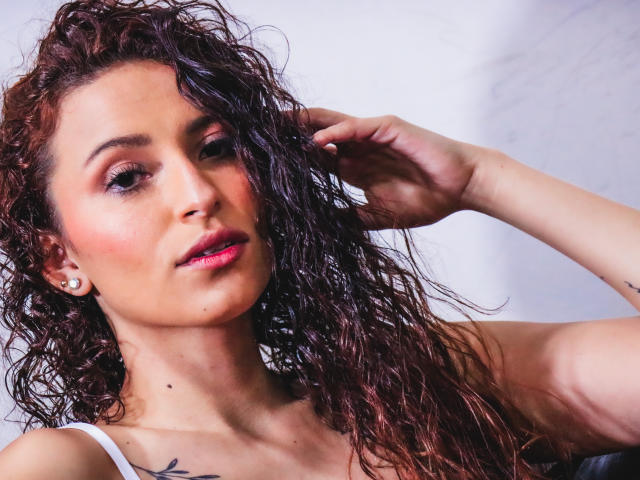 AnnaMary tiene el pelo castaño rizado y húmedo, lápiz labial rosa y un tatuaje en el hombro derecho. Está mirando hacia un lado, con la mano en el pelo, mostrando sus pechos..