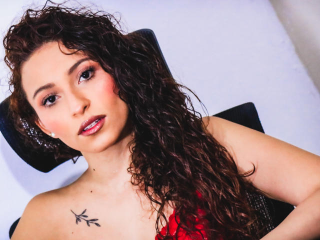 AnnaMary, com cabelos castanhos cacheados, batom vermelho e uma pequena tatuagem no ombro direito, vestindo uma blusa vermelha, sentada com um apoio de cabeça preto, olhando para a câmera com um leve sorriso..