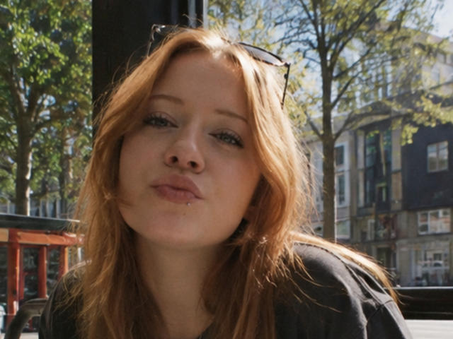 SerafinaDinwiddie has a nose piercing, pouty lips, and slightly wavy blonde hair, wearing a black top, with sunglasses on her head, in a sunny urban setting.