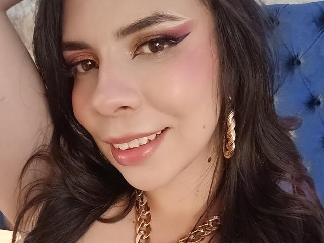 VioletaStone has shiny brown eyes, pink eyeliner, and a cute smile with glossy pink lips. She's wearing gold earrings and a chunky gold chain, with dark wavy hair.