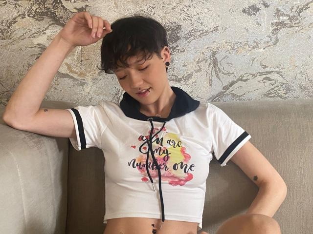 LissyLuxe with short dark hair, small black ear holes, white crop top, black hood, "I am my own water" text, sitting on gray couch, arm raised, midriff exposed.