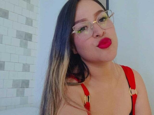 LiaDiane with long, straight, blonde-tipped black hair, wearing red suspenders and red lipstick, pouting with green eyeshadow and clear glasses, showing her tits.