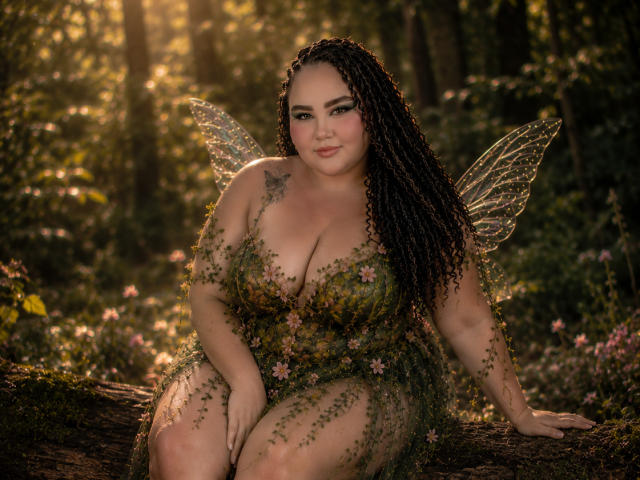 HurrenMatt mit lockigem, dunklem Haar, Feenflügeln und einem grünen, blumigen Kleid, sitzt auf einem Baumstamm, zeigt große Brüste, ein Tattoo an ihrer linken Schulter und ein süßes Lächeln..