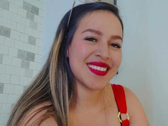 LiaDiane, avec du rouge à lèvres, souriante, portant une robe rouge avec des bretelles, a de longs cheveux bruns avec des mèches blondes, et tient des lunettes de soleil sur sa tête..