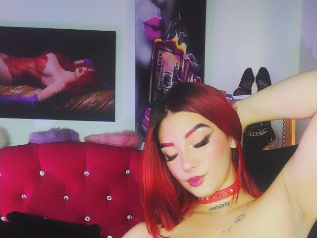 BriielaEva with red hair, wearing a red choker, has her arm up, showing her armpit, and has bold eyeliner, pink lipstick, and a nose ring. Boobs are visible.