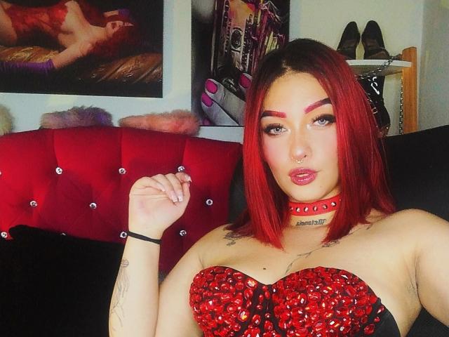 BriielaEva with red lipstick, red-studded choker, and red sequined top, showing off her tits and tattoos, with red and black hair, and a black bracelet.