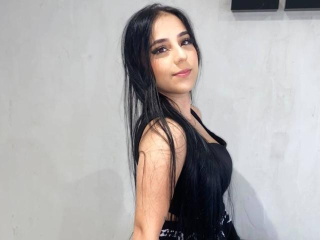 StefanyDelly a de longs cheveux noirs, une peau brun clair, et porte un haut noir qui révèle ses petites seins et un sourire discret. Elle regarde par-dessus son épaule..