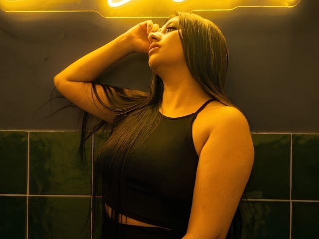 CatalinaRivero with long black hair, wearing a black crop top, standing with one arm raised, looking to the side, under warm yellow lighting. Her tits are visible.