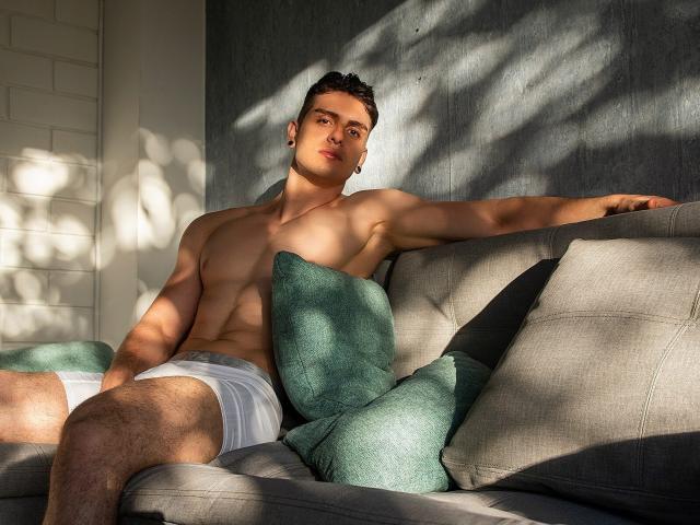 Aaron Kwok, senza camicia, con un corpo muscoloso, tonico e dalla pelle chiara, indossa biancheria intima bianca, ci sono cuscini verdi e un divano grigio, è appoggiato all'indietro, con un braccio disteso, in ombra a causa della luce del sole, ha dei piercing alle orecchie e i capelli sono corti e ricci..