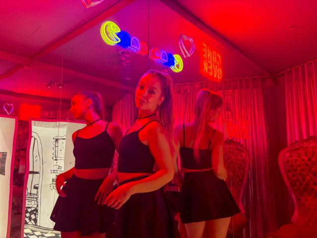 Trois jeunes femmes, les cheveux longs, portant des tops courts noirs et des jupes, debout dans une pièce éclairée en rouge, avec des néons au-dessus, des reflets montrant leurs fesses..