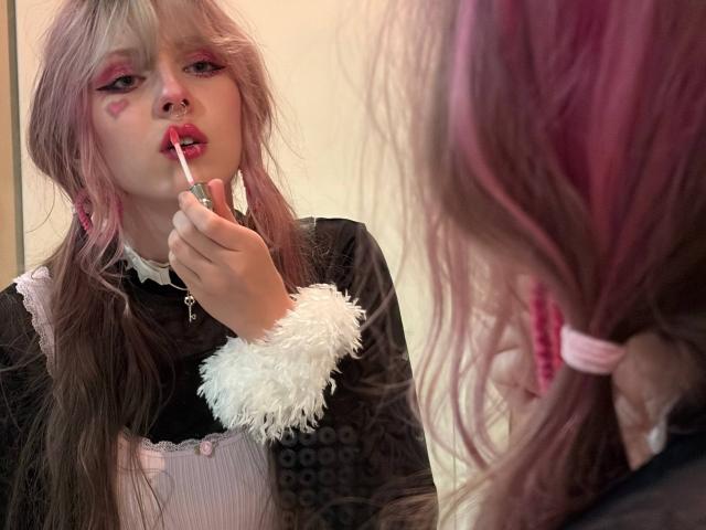 Louella Cardenal, con i capelli rosa, indossa una maglietta nera e bianca e si sta applicando rossetto rosa sulle labbra con un bastoncino di rossetto rosso, come si vede riflesso in uno specchio..