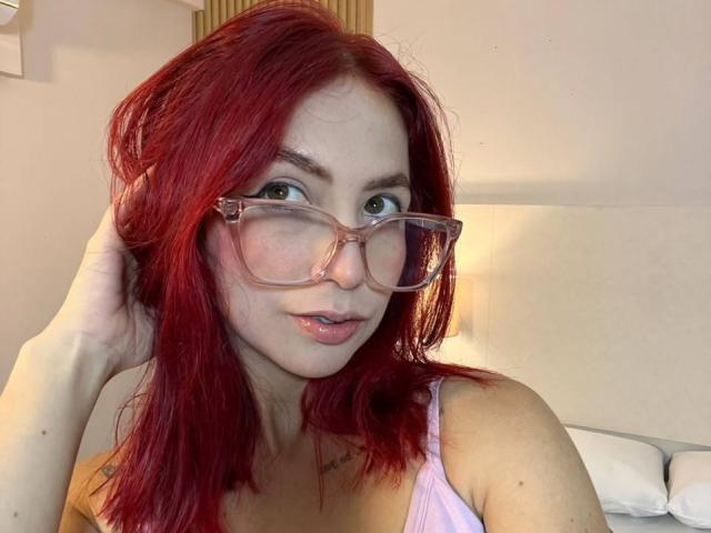 MiaaRouge, con i capelli rossi, indossa occhiali rosa e una canottiera rosa, ha una mano tra i capelli, guarda la fotocamera con gli occhi verdi e le labbra leggermente dischiuse..