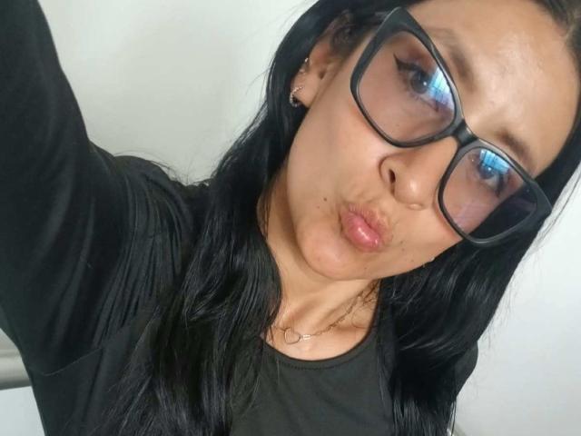 ZarayFeet with pouty lips, black glasses, and long black hair, wearing a black shirt, has a small beauty mark under her right eye, and a heart necklace.