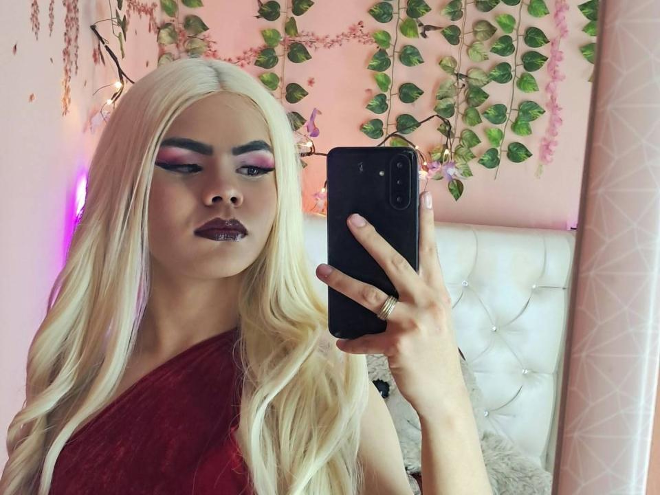 DanySky with long blonde hair, red one-shoulder dress, dark pink eyeshadow, purple lipstick, holding phone, taking selfie, wearing ring, in pink-lit room, vines in background.