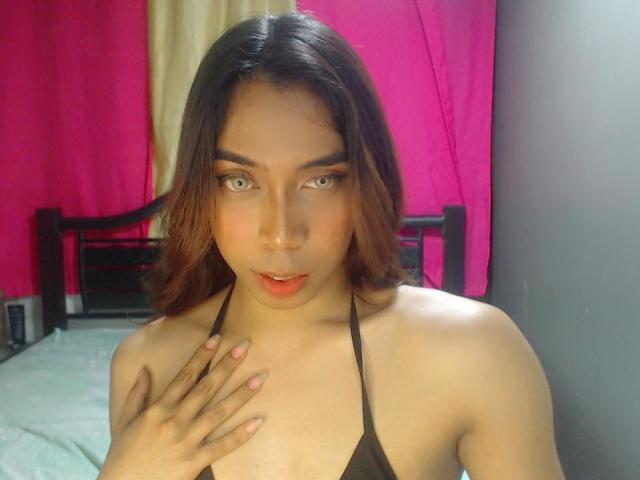 SophiaBrown: Uma mulher de cabelos castanhos, pele clara e olhos cinzentos, vestindo uma blusa preta de alças, com a mão no peito, olhando para a câmera com a boca ligeiramente aberta, fundo rosa e bege..