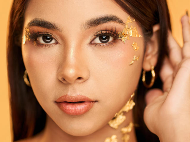 NathalieAndLuna's face with golden leaf makeup on her right cheek and ear, brown eyes, long brown hair, orange lipstick, gold hoop earrings, and a hand touching her hair.