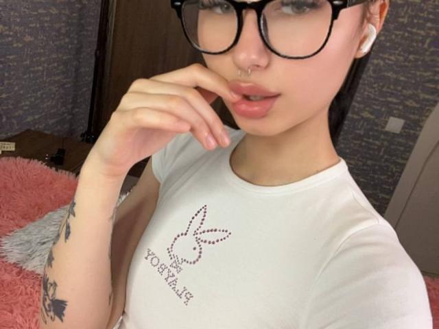 GlitchDoll with glasses, pale skin, and a nose ring, touching her lips with her finger, wearing a white shirt, showing a tattoo on her arm, and pink lips.