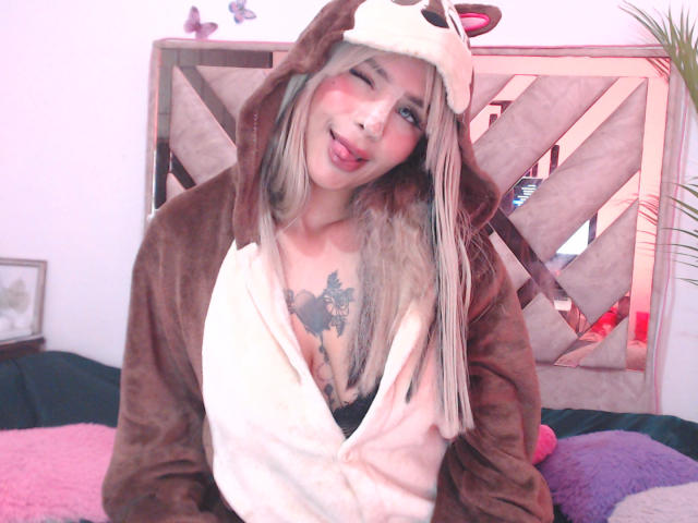 LuluDeVine with blonde hair, pink blush, and tongue out, wearing a brown and white furry hood, exposing black bra and chest tattoo, in a cozy room.