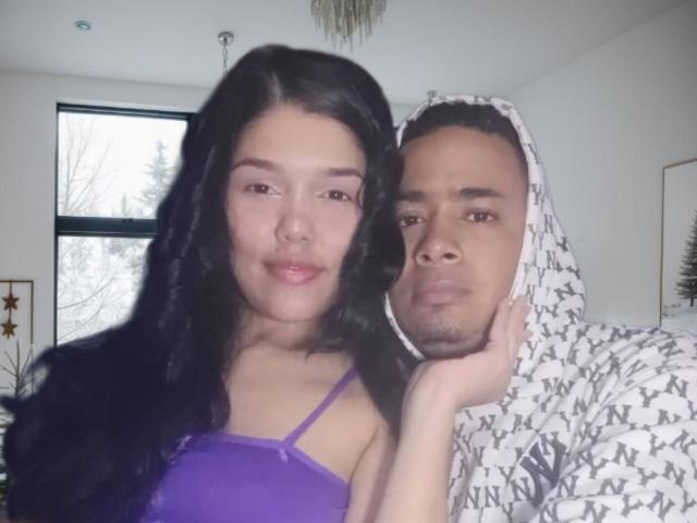 CoupLatina with long black hair in a purple top and a brown-skinned dude in a white patterned hoodie, both with serious faces, standing closely together in a room.