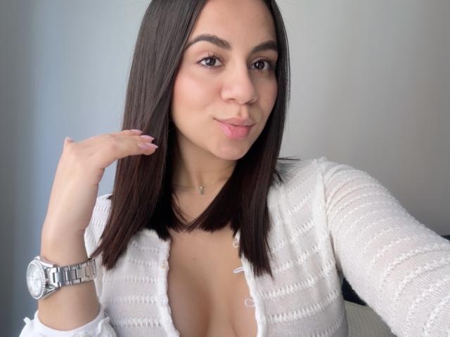 EmaRusso with straight brown hair, wearing a white, ribbed, open-front top, showing her cleavage, pink lipstick, and a silver watch, puckering her lips.