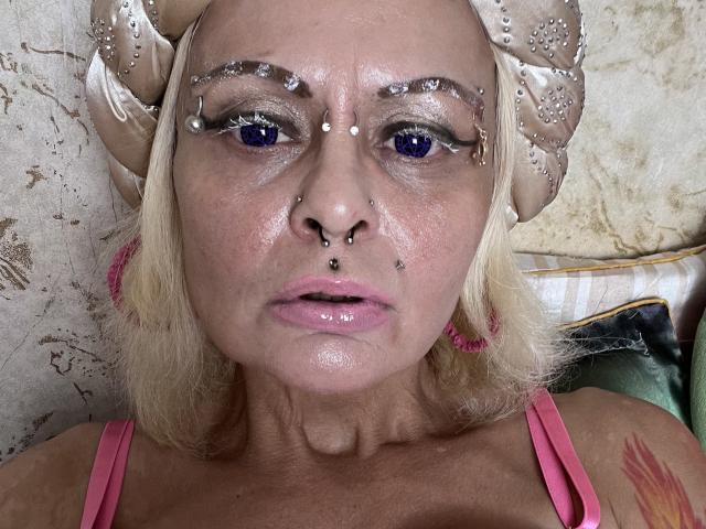 Icecandyshokolade has blue eyes, a nose ring, and a septum piercing. She's wearing a beige headband with ribbons, has glitter on her eyelids, and pink lipstick. Her tits are visible.