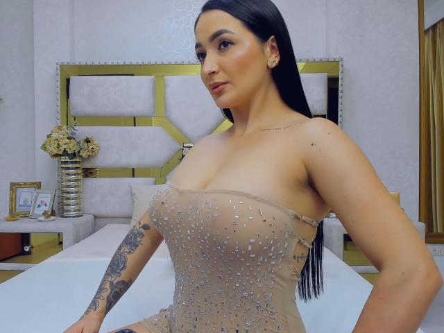 PamelaLiss heeft grote borsten, een getatoeëerde arm, en draagt een doorschijnende, met juwelen versierde jurk, met haar zwarte haar los, terwijl ze op een bed in een slaapkamer zit..