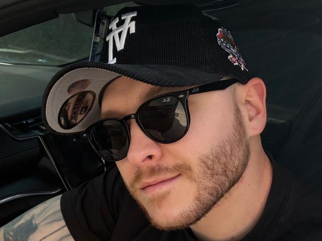 AdamMeyer with a beard, wearing a black cap with a logo, black sunglasses, and a black shirt, sitting in a car, looking slightly to the side.