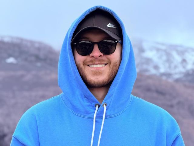 AdamMeyer with a blue hoodie, black cap, and dark sunglasses, smiling with a beard, standing outdoors, wearing a Nike cap, showing a medium build, and a happy expression.