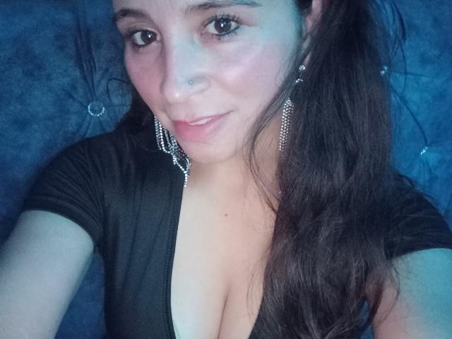 Crisht a de longs cheveux bruns, un piercing au nez, et porte un haut noir en V qui dévoile ses seins. Elle sourit, ses yeux sont bleus, elle porte des boucles d'oreilles pendantes et a un joli visage..