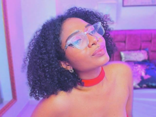 MeganWilliams with curly black hair, wearing clear glasses and a red choker, has a nose ring and is pouting with glossy lips. Boobs not visible, focus on upper body.