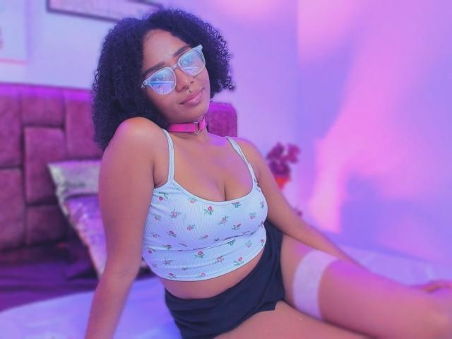 MeganWilliams with curly black hair, wearing glasses, a pink choker, a light blue tank top with small patterns, black shorts, and white thigh-high socks, sitting on a bed.