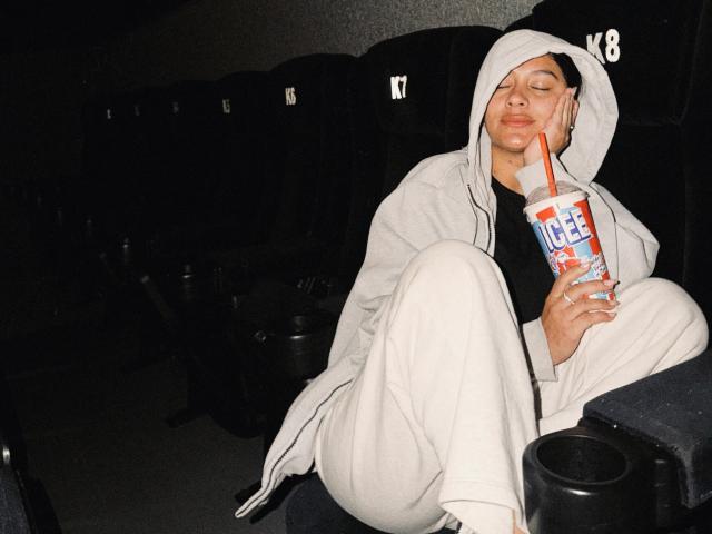 ThaliaAres, seduta in un teatro buio, indossa una felpa bianca e dei pantaloni, tiene in mano un ICEE con una cannuccia rossa, ha gli occhi chiusi, sorride, ha una mano sul viso, appare rilassata..