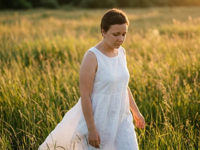 GoldenHaze, short brown hair, white sleeveless dress, looking down, standing in tall green grass, sunlight, gentle expression, soft light, slight breeze, subtle shadow, natural setting.