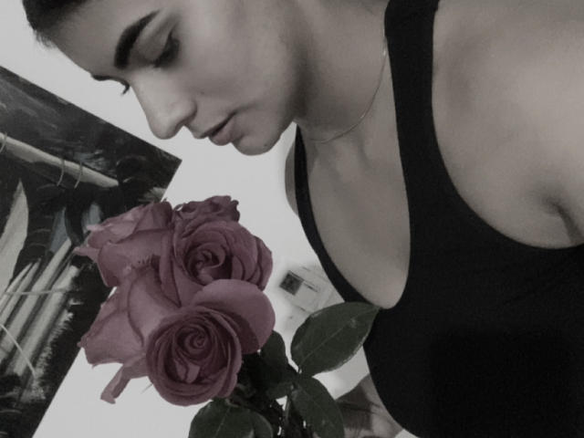 AlaraKey with dark hair, black tank top, looking down at pink roses in a photo. Her tits are visible, and she's wearing a thin necklace. Black-and-white photo.