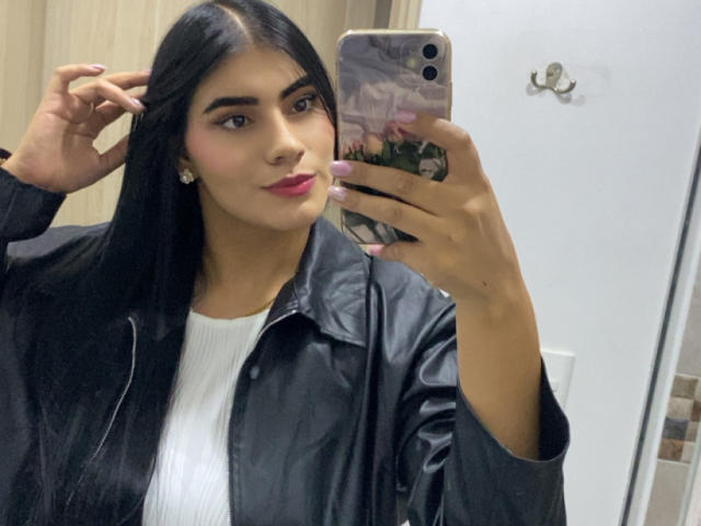 AlaraKey with long black hair, wearing a black leather jacket and white ribbed shirt, taking a mirror selfie with a pink phone case, pink lipstick, and pink nails.