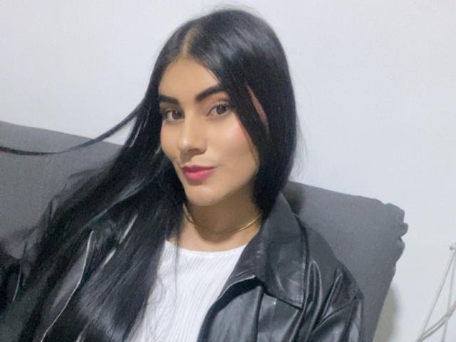 AlaraKey has long black hair, brown eyes, and pink lipstick. She's wearing a black leather jacket over a white ribbed shirt. Her ass is not visible.
