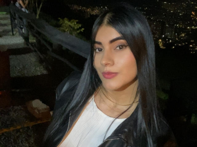 AlaraKey has long black hair, dark eyebrows, pink lipstick, and wears a black leather jacket over a white top. Her ass is not visible.