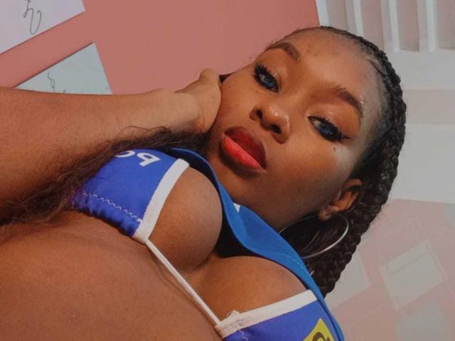 Shaaron's face is close to a hand, wearing a blue sports bra that reveals her large boobs. She has red lipstick, braided hair, and hoop earrings. Her eyes are half-open.