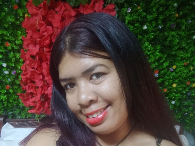 MariahSquirting69 with dark brown hair, red lipstick, smiling, showing her shiny face and wearing a black necklace. She has cute tits and a small nose. Green background.