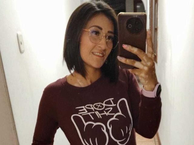 SharissWilliam with short black hair, glasses, and a maroon shirt, holding a phone in a mirror selfie, showing her tits and a nose ring, smiling.