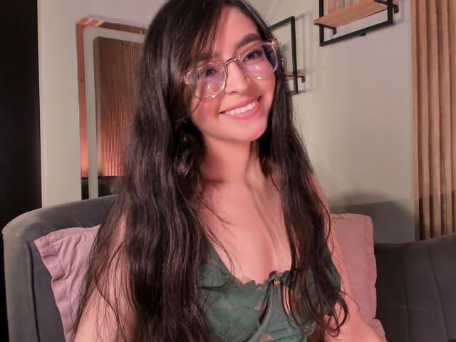 EvaMcAllister with long dark hair, wearing glasses and a green lace bralette, smiling at the camera, showing her tits. She's sitting on a grey couch with pink pillows.