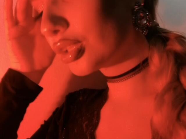 ElzaKomx's face, red-lit, with tongue out, slightly glossy lips, black choker, wavy brown hair, hand near ear, wearing black top, earrings, and a sexy, seductive expression.